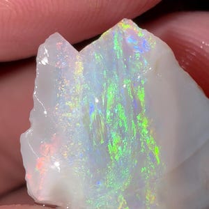 May include: A raw, white opal gemstone featuring a vibrant play-of-color with flashes of bright green, blue, and orange light across its irregular, translucent surface. The stone has a jagged, unpolished edge and a smooth, glossy texture.