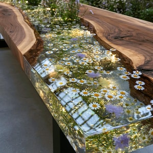 Peut inclure: Une longue table rectangulaire composée de deux plateaux en bois de noyer à bords naturels, séparés par un canal central en résine transparente. La résine contient des marguerites blanches et des petites fleurs violettes, sur pieds en métal noir.