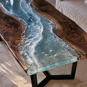 May include: A rectangular coffee table featuring two live-edge dark brown wood slabs separated by a central river of translucent blue resin. The resin mimics ocean waves with white foam detailing and small embedded gold-toned accents on black legs.
