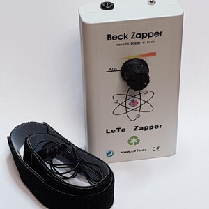 Könnte beinhalten: Ein weißes, rechteckiges elektronisches Gerät mit der Aufschrift Beck Zapper Nach Dr. Robert C. Beck und LeTe Zapper, ausgestattet mit einem schwarzen Drehknopf, einer Atomgrafik und einer Webadresse. Ein schwarzes Stoffband mit Kabel liegt daneben.