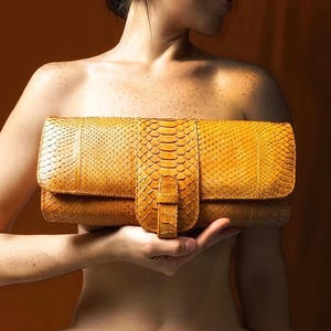 May include: A rectangular mustard yellow clutch bag featuring a textured snakeskin pattern. The bag has a horizontal flap closure secured by a vertical strap with a matching snakeskin finish. The surface displays a consistent scale-like texture.