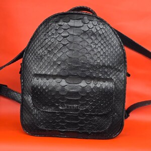 Puede incluir: Una mochila negra con un patrón de piel de serpiente texturizada en todo el exterior. La bolsa incluye un bolsillo frontal con cierre de solapa y el texto EikanReptile impreso en letras blancas pequeñas sobre la solapa del bolsillo frontal.