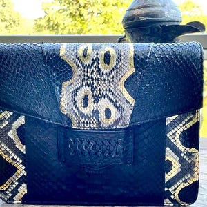 May include: A rectangular handbag featuring a black snakeskin texture with contrasting panels of grey, black, and gold snakeskin patterns. The front flap includes a central vertical strip of patterned scales and a horizontal woven leather detail.