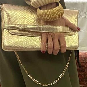 May include: A metallic gold clutch bag with a textured snakeskin pattern, featuring a horizontal strap across the front and a thin gold chain shoulder strap. Two thick, ribbed, cream and tan colored bangles are worn on the wrist.