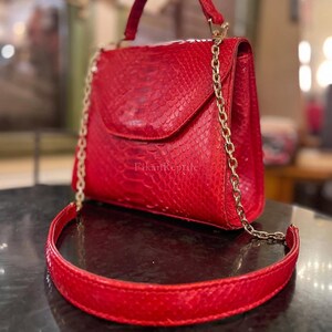 Puede incluir: Un bolso de mano rojo brillante con un patrón texturizado de piel de serpiente, que cuenta con un asa superior, una solapa de cierre y una correa de cadena dorada con una hombrera de cuero rojo. El texto FikanReptile está impreso en la solapa.