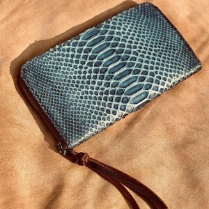 May include: A rectangular wallet featuring a blue and black snakeskin-patterned exterior with a textured, scaled surface. The wallet is secured with a dark zipper closure and includes a brown leather pull tab with two long, thin leather strips.