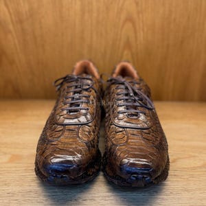 May include: A pair of brown sneakers featuring a textured, scale-like pattern across the upper surface. The shoes have dark brown laces, a rounded toe box, and a low-top silhouette. The text "FikanReptile" is visible on the tongue of the shoes.