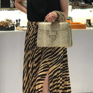 May include: A structured handbag featuring a metallic gold snakeskin-textured finish, a top handle, and a flap closure with a central tab detail. The bag includes a gold-tone chain strap and is held by a top handle, showcasing a reptilian pattern.