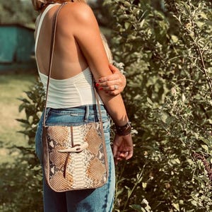 Peut inclure: Un sac à bandoulière avec un motif en peau de serpent dans des tons marron, beige et crème. Le sac possède une longue bandoulière fine et une fermeture à bascule centrale faite d'un matériau incurvé clair rappelant un bois ou un os.
