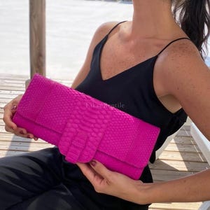 May include: A vibrant hot pink clutch bag featuring a textured snakeskin pattern and a central vertical strap closure. The rectangular accessory has a flap top design and displays the text FikanReptile printed in the center of the front panel.