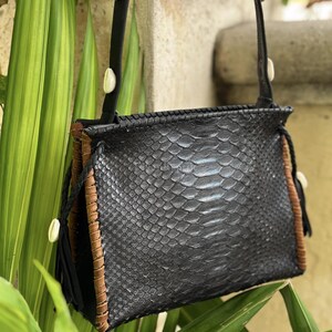 Puede incluir: Un bolso de hombro negro con textura de piel de serpiente, paneles laterales de cuero marrón y detalles de costura. El bolso incluye una correa negra adornada con conchas de cauri blancas y borlas de cuero negro en los laterales.