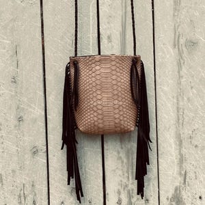Peut inclure: Un sac bandoulière beige avec un motif texturé en peau de serpent sur le corps principal. Le sac présente de longues franges noires suspendues sur les deux côtés et une fine bandoulière noire fixée aux coins supérieurs de la pochette.