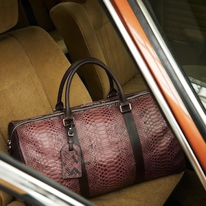 May include: A dark burgundy duffel bag with a textured snakeskin pattern, featuring dark brown leather handles, a matching luggage tag, and a silver-toned zipper closure. The bag is positioned on a tan fabric car seat with vertical stitching.