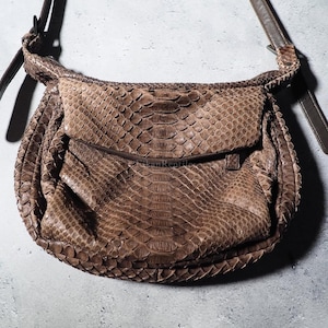 Puede incluir: Un bolso de hombro marrón con un patrón de piel de serpiente texturizado en todo el exterior. El bolso cuenta con un cierre de solapa curvado, un compartimento principal estructurado y una correa larga, fina y ajustable en los laterales.