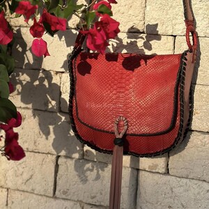 May include: A red snakeskin-textured crossbody bag with a rounded flap, black stitched edges, and a long, thin, light brown leather tassel hanging from a central braided loop. The bag features a shoulder strap attached with circular rings.