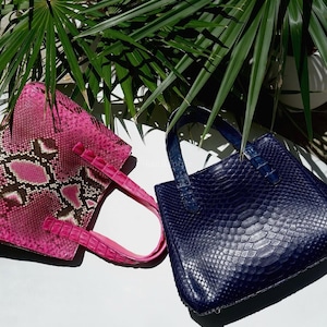 May include: Two structured handbags with a textured snakeskin pattern. One bag is vibrant pink with dark brown and white diamond-shaped accents, while the other is a solid deep navy blue. Both feature matching top handles and a rectangular shape.