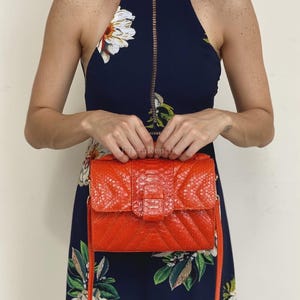 May include: A bright orange rectangular handbag with a textured snakeskin-like pattern and a chevron-stitched design. The bag features a top handle, a front flap closure with a central tab, and a thin shoulder strap. The surface has a glossy finish.