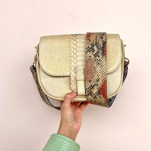 Peut inclure: Un sac bandoulière de couleur crème avec un motif texturé en peau de serpent. Le sac présente une fermeture à rabat avec un détail de sangle verticale et une large bandoulière amovible affichant un imprimé serpent multicolore rouge, noir et beige.