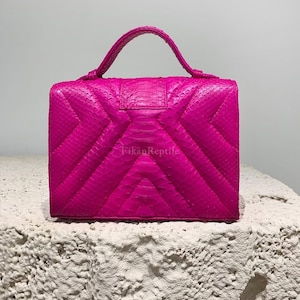 Puede incluir: Un bolso de mano rosa intenso con un patrón texturizado de piel de serpiente y una forma rectangular estructurada. El bolso cuenta con un asa superior y un cierre de solapa con un diseño de costuras en chevrón. El texto FikanReptile está impreso.