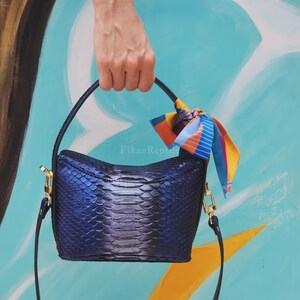 Op de afbeelding: Een donkerblauwe handtas met een gestructureerd slangenleerpatroon en een metallic glans. De tas heeft een handvat, een afneembare schouderriem met goudkleurige hardware en een kleurrijke zijden sjaal aan de zijkant. De tekst FikanReptile is zichtbaar.