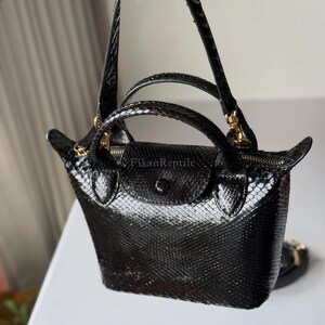 May include: A black handbag with a glossy snakeskin texture, featuring a top flap with a snap closure, two short handles, and a longer shoulder strap. The text FikanReptile is printed on the front flap. The bag includes gold-toned hardware.