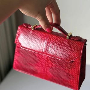 May include: A bright red structured handbag featuring a textured snakeskin-like pattern across the exterior. The bag includes a top handle attached with gold-toned metal hardware and a flap closure that covers the front compartment of the bag.