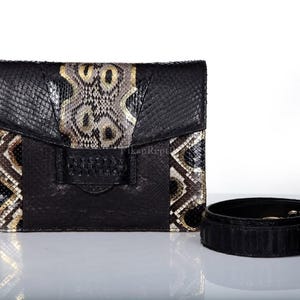 May include: A rectangular black handbag featuring a textured finish and a central vertical panel with a grey, black, and gold snakeskin pattern. The bag includes a matching black detachable shoulder strap coiled beside it on a white surface.