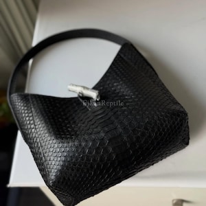 May include: A black handbag with a textured, scale-like pattern across the exterior. The bag features a single shoulder strap and a silver-toned toggle closure at the top centre. The text "RikanReptile" is printed in small letters near the closure.
