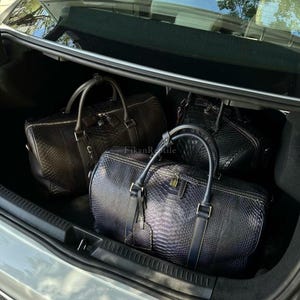 Puede incluir: Tres bolsas de viaje con un patrón de piel de serpiente texturizado están dispuestas en el maletero de un coche. Una bolsa es azul oscuro, mientras que las otras dos son marrón oscuro. Cada bolsa tiene dos asas, cremallera y una etiqueta.