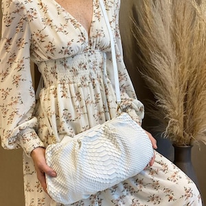 Puede incluir: Un bolso de mano blanco con un patrón texturizado de escamas y una correa fina para el hombro, combinado con un vestido de manga larga color crema con escote en V, cintura fruncida y un delicado estampado floral en tonos marrón y rosa.