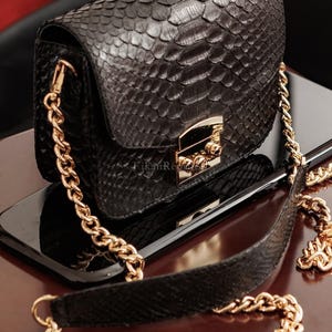 May include: A black handbag with a textured snakeskin pattern, featuring a gold-tone metal chain strap and a matching gold-tone rectangular clasp closure on the front flap. The bag has a structured rectangular shape with visible scale details.