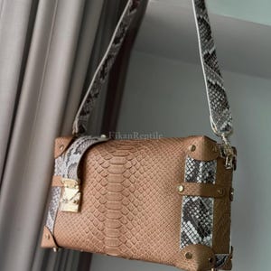 Puede incluir: Un bolso rectangular con cuerpo de textura de piel de serpiente color canela, paneles laterales en contraste con estampado de piel de serpiente gris y blanco y una correa a juego. Incluye refuerzos de esquina y un cierre en tono dorado.