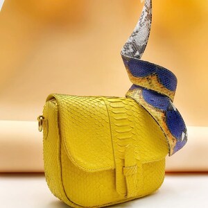 May include: A bright yellow crossbody bag with a textured snakeskin pattern. The bag features a flap closure with a vertical strap detail and a wide, detachable shoulder strap that displays a multi-coloured snakeskin print in blue, yellow, and grey.