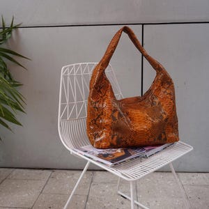 Op de afbeelding: Een hobo-handtas met een gestructureerd, oranje en bruin slangenleerpatroon. De tas heeft een enkele geïntegreerde schouderband en een slappe, ongestructureerde vorm met een brede opening aan de bovenkant en een taps toelopende onderkant.