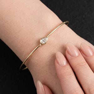 May include: A thin gold bangle bracelet featuring a single pear-shaped clear gemstone set in a bezel setting at the center. The bracelet has a smooth, polished finish and a minimalist design that encircles the wrist with a delicate, slender band.