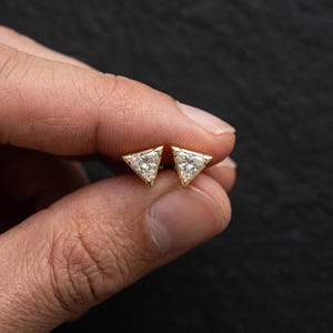 Op de afbeelding: Een paar oorknopjes met driehoekig geslepen heldere stenen gezet in geelgoud. Elke oorbel heeft een puntige driezijdige vorm met een gefacetteerd oppervlak dat licht reflecteert, vastgehouden door een gouden metalen frame met kleine pootjes.