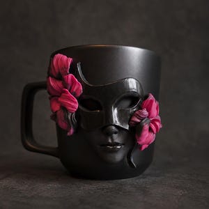 Op de afbeelding: Een matzwarte keramische mok met een gebeeldhouwd zwart carnavalsmasker in het midden. Het masker wordt aan beide kanten geflankeerd door levendige magenta en donkerpaarse bloemaccenten die in het oppervlak van de mok zijn verwerkt.