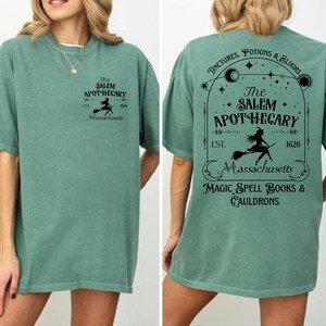 a woman wearing a green t-shirt with a design that includes text and graphics related to a witch-themed business called "The Salem Apothecary".