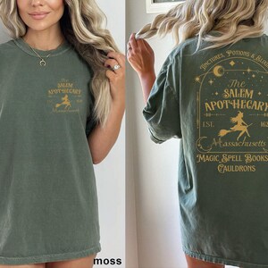A woman wearing a green t-shirt with text and graphics related to the Salem Apothecary.