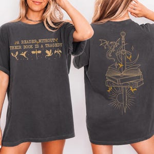 May include: A charcoal gray short-sleeve t-shirt featuring gold graphic prints. The front displays the text A READER WITHOUT THEIR BOOK IS A TRAGEDY with small dragon silhouettes. The back features a large illustration of a sword, books, and dragons.