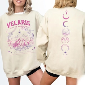 May include: A cream-colored crewneck sweatshirt featuring a pink and purple graphic print. The front displays the text VELARIS CITY OF STARLIGHT above a mountain landscape and floral frame. The back shows a vertical sequence of moon phases.