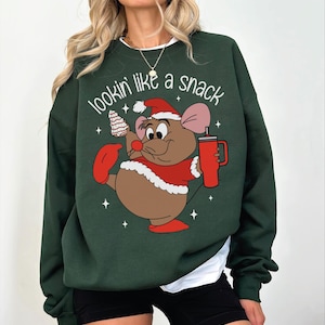 May include: A dark green crewneck sweatshirt featuring a graphic of a brown cartoon mouse wearing a red Santa hat and coat. The mouse holds a striped treat and a red tumbler. The text "lookin' like a snack" is printed in white above the mouse.
