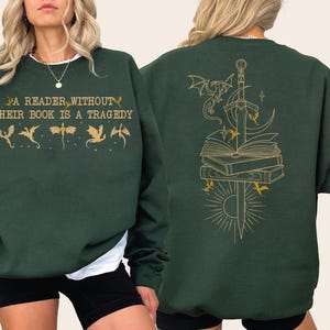 May include: A dark green crewneck sweatshirt featuring gold-colored graphics. The front displays the text A READER WITHOUT THEIR BOOK IS A TRAGEDY with small dragon silhouettes. The back shows a large illustration of a sword, books, and dragons.