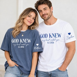 May include: A blue t-shirt with white text reading GOD KNEW my heart needed you Est.2026 and a white heart with the name William on the sleeve, and a white t-shirt with blue text reading GOD KNEW my heart needed you Est.2026 and a blue heart with Sophia.