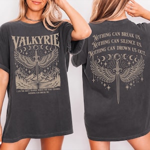 May include: A charcoal gray short-sleeve t-shirt featuring a beige graphic of a winged sword, mountains, and moon phases. The front text reads VALKYRIE and I AM THE ROCK AGAINST WHICH THE SURF CRASHES. NOTHING CAN BREAK ME. The back text reads NOTHING CAN BREAK US. NOTHING CAN SILENCE US. NOTHING CAN DROWN US OUT.