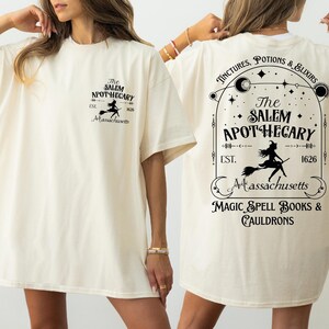 a woman wearing a white t-shirt with text and graphics related to a witch-themed business.