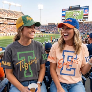 May include: A charcoal gray t-shirt with a green block letter T, the word Tigers in script, and the text NAME and 12 on the sleeves, paired with a green and yellow baseball cap. A peach t-shirt with a blue block letter E, the word Eagles in script, and the text NAME and 17 on the sleeves, paired with a blue and orange baseball cap.