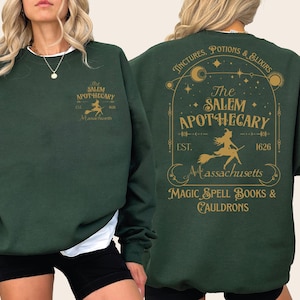 May include: A dark green long-sleeve sweatshirt featuring gold graphic prints. The front displays a small witch on a broomstick with the text The Salem Apothecary Est 1626 Massachusetts. The back features a large ornate arch design with celestial motifs, the text The Salem Apothecary, and the words Magic Spell Books & Cauldrons.