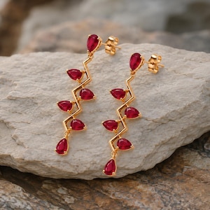 Puede incluir: Un par de pendientes colgantes dorados con una estructura metálica en zigzag adornada con varias piedras preciosas rojas en forma de pera. Cada pendiente tiene un cierre de presión y un diseño vertical que alterna las piedras a lo largo del marco.