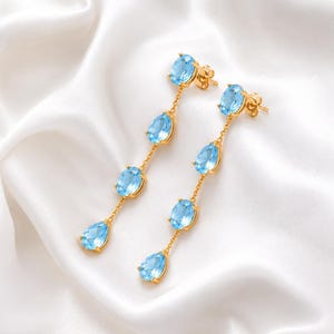 Puede incluir: Un par de pendientes colgantes de oro, cada uno con tres piedras preciosas de color azul claro en forma ovalada dispuestas verticalmente en cadena. Las piedras están engastadas en metal dorado, con la piedra superior sirviendo como poste.